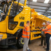 Large yellow gritter with Via logos in exhibitions hall. The driver door is open and two gritter drivers in orange jackets are showing a councillor around. He wears the chairman's chain