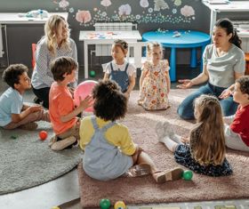 A classroom with young children sat down on the floor with two female teachers present. 